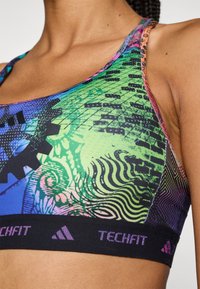 Multicolorde sportbeha met een levendig grafisch ontwerp en geometrische patronen, gemaakt van rekbaar materiaal. Zwarte band met het "TECHFIT" logo.