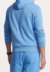 Person som bär en ljusblå huvtröja och matchande sweatpants, sedd bakifrån, med höger arm avslappnad och en klocka på vänster handled.