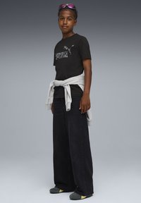 Musta t-paita, jossa on harmaa Puma-logo, harmaa huppari sidottuna vyötärölle, leveät mustat farkut ja harmaat kengät keltaisilla yksityiskohdilla.