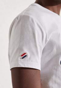 Bílá bavlněná tričko s krátkým rukávem a malým látkovým logem na rukávu v červené, modré a bílé barvě. Měkká textura.