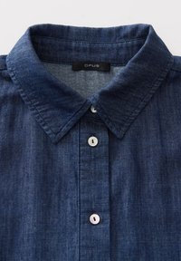 Chemise en denim bleu foncé à boutons avec col pointu et boutons nacrés, affichant l'étiquette de la marque "OPUS" à l'intérieur du col.