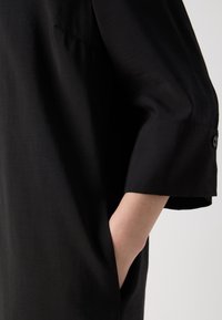 Personne portant une chemise noire à manches longues avec un poignet boutonné, main partiellement insérée dans une poche latérale sur un fond clair uni.