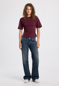 Maroon short-sleeve top with ruching at the waist, paired with wide-leg blue jeans and white sneakers, standing against a plain background.