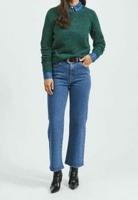 Suéter de punto verde, camisa de mezclilla azul debajo, jeans azul de cintura alta y botines negros de punta afilada. Materiales texturizados, fit relajado.