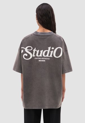 Donna con lunghi capelli scuri che indossa una maglietta grigia oversize con testo bianco "Studio Crafting Aesthetics WRSTBHVR" sulla schiena.