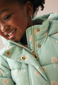 Hellgrüne Steppjacke mit floralem Muster, goldenen Details, einem Reißverschluss und Druckknöpfen. Sie hat einen hohen Kragen und eine weiche Textur.