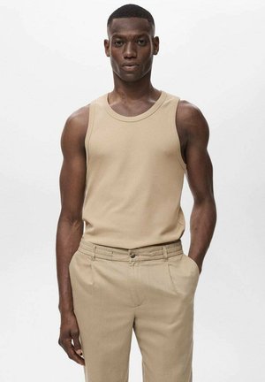 Débardeur beige à col rond, porté avec un pantalon beige clair, de texture lisse. Le mannequin se tient debout avec les mains dans les poches.
