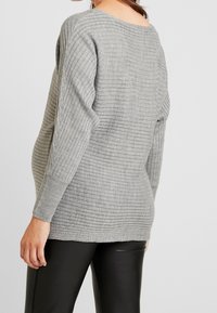 Kobieta ubrana w szary, prążkowany sweter z długimi rękawami i czarne skórzane spodnie, pokazana od tyłu na jednolitym tle.