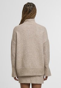 Beige, kötött pulóver magas nyakkal, laza szabású és bordás mandzsettával. Puha textúra és finom színváltozatok futnak végig az anyagon.