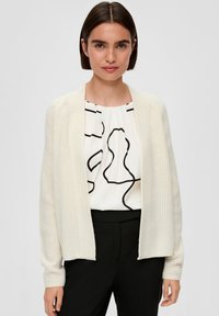 Crèmekleurige gebreide cardigan met ribbelstructuur, open voorkant en lange mouwen. Gekozen over een witte blouse met een zwart abstract patroon.