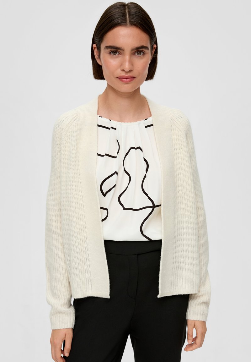 Crèmekleurige gebreide cardigan met ribbelstructuur, open voorkant en lange mouwen. Gekozen over een witte blouse met een zwart abstract patroon.