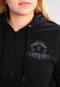 Felpa nera con zip realizzata in tessuto morbido, caratterizzata da una stampa grafica con la scritta "NEW YORK BY HEART & SOUL" in bianco e accenti argentati.