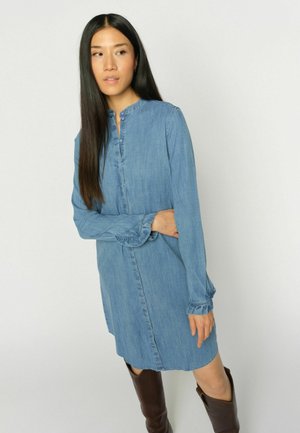 Hosszú sötét hajú nő, aki kék farmer hosszú ujjú ruhát és barna, térdig érő csizmát visel, egyszínű világos háttér előtt áll.