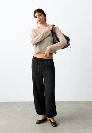 Mlajša ženska v širokih črnih hlačah, bež kratki dolgi majici z dolgimi rokavi in rjavih nizkih čevljih, s črno torbo čez ramo ob beli steni.