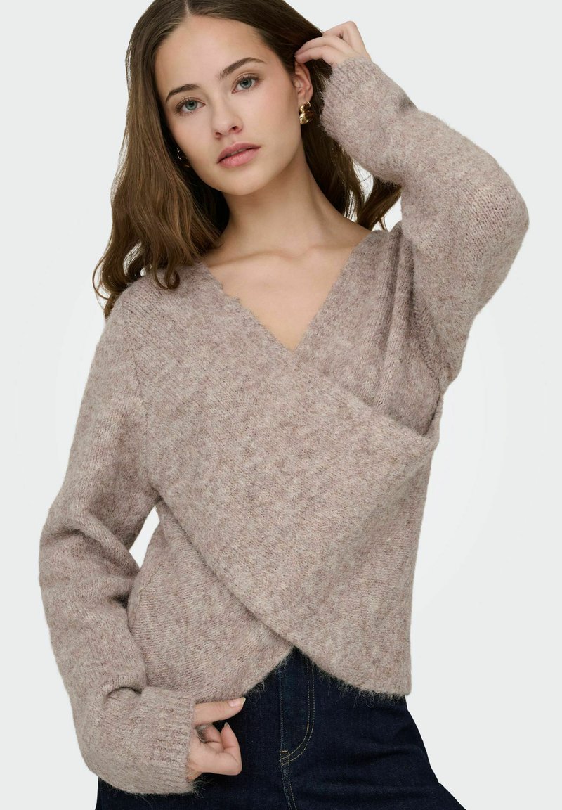 Pull en maille douce couleur brun clair avec un col en V profond, une coupe ample et des manches longues ; présente un design croisé avec une finition texturée.