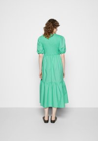 Vestido de algodão verde com corpete franzido, mangas bufantes e saia em camadas, usado com sandálias pretas de slide. Vista traseira mostrada.