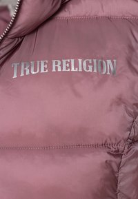 Růžová nafouknutá bunda se hladkým, lesklým povrchem, s vyraženým textem "TRUE RELIGION" v stříbrné barvě na oblasti hrudníku.
