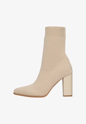 Beige enkellaars met een ribgebreide bovenkant, spitse neus en een blokhak. Heeft een gladde textuur en een naadloos ontwerp met een contrasterende zool.