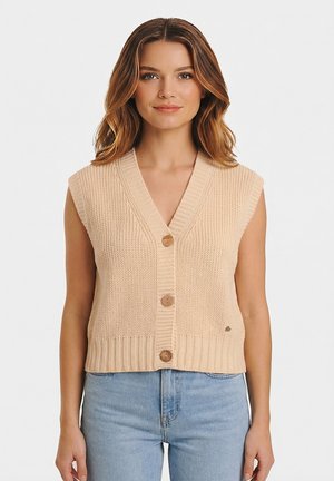 Donna con capelli castani ondulati indossa un gilet lavorato a maglia beige senza maniche con bottoni e jeans azzurri a vita alta, in piedi davanti a uno sfondo semplice.