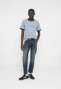 Ανοιχτό μπλε μπλουζάκι με ναυτικά στοιχεία και τη λέξη «ORIGINALS», συνδυασμένο με σκούρο μπλε τζιν και μαύρα αθλητικά παπούτσια με λευκές ρίγες.