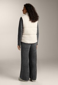 Frau mit lockigem Haar, die eine weiße Sherpa-Weste über dunkelgraßen, weit geschnittenen Strickhosen und passendem Pullover trägt, steht vor einem neutralen Hintergrund.