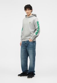 Young man wearing grey hoodie with green "BARROW" text on sleeves, blue jeans, and black sneakers, standing with one hand in pocket.