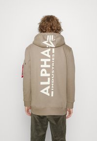 Khaki kapuca s kapuco s velikim belim printom "ALPHA INDUSTRIES" na leđima, rebrasti manžeti i teksturiran finis. Crvena oznaka na rukavu.