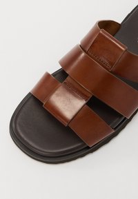 Sandale en cuir marron avec trois larges brides croisées, semelle foncée et finition lisse. Le dessus présente un discreet détail de couture.
