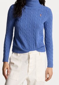 Sininen palmikkoneulepusero korkealla kauluksella, valmistettu pehmeästä materiaalista, jossa on resorit hihansuissa ja helmassa sekä pieni logoyksityiskohta rinnassa.