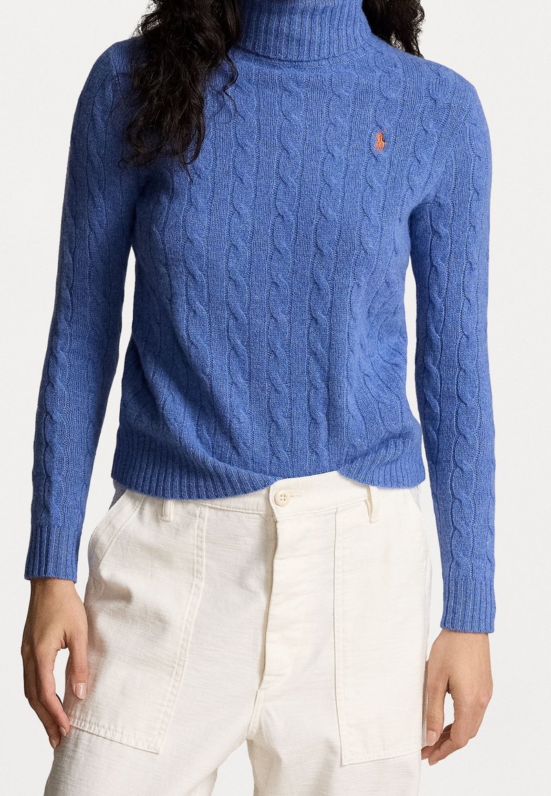 Sininen palmikkoneulepusero korkealla kauluksella, valmistettu pehmeästä materiaalista, jossa on resorit hihansuissa ja helmassa sekä pieni logoyksityiskohta rinnassa.