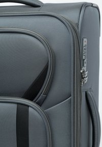 Valise à roulettes grise avec un tissu texturé, plusieurs compartiments zippés, un verrou à combinaison et deux poignées pour un transport facile.