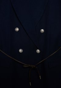 Blazer doppiopetto blu navy con bottoni argentati, cintura marrone in camoscio e tessuto strutturato. Presenta un design elegante e tasche laterali.