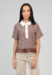 Jeune femme portant un haut en tweed multicolore à manches courtes avec un col blanc, des boutons décoratifs, un ourlet frangé et un pantalon beige, se tenant devant un fond gris.