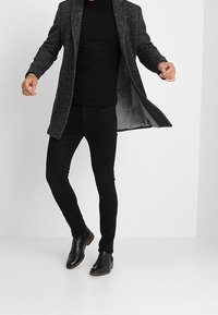Persoon in een donkergrijze coat, zwarte coltrui, zwarte skinny broek en zwarte leren laarzen, posed met open coat tegen een witte achtergrond.