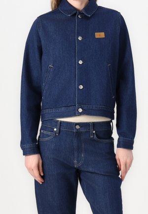 Person iført en mørkeblå denimjakke med sølvknapper og matchende denimjeans mod en ensartet lys baggrund.