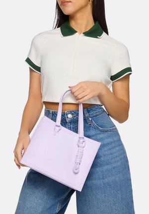 Sac fourre-tout en cuir lavande avec une forme rectangulaire structurée, détail de logo embossé et poignées courtes. Porté avec un haut blanc et un jean bleu.