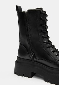 Fekete bőr bokacsizma masszív talppal, fűzős elejével és oldalsó cipzárral. Simább felülettel rendelkezik, és texturált talpa van a tapadás érdekében.