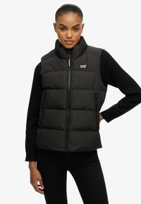 Chaleco acolchado negro con cuello alto, cierre de cremallera y bolsillos laterales. Presenta un diseño acolchado texturizado y un logo en el pecho izquierdo.