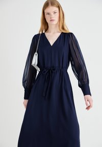 Navy dress with sheer puff sleeves, V-neck, and a drawstring waist. Smooth fabric with a flowy silhouette. Silver handbag accent.
