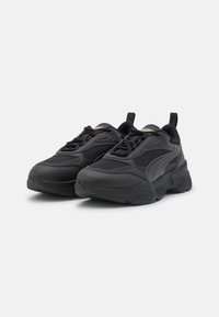 Black athletic sneakers with a textured mesh and synthetic upper, featuring a thick sole and a rear pull tab for easy wear. Gold accents present.