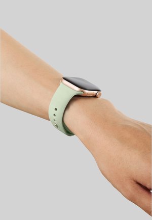 Pols met een roségouden smartwatch met een lichtgroene siliconen band op een lichte huidtint, tegen een effen grijze achtergrond.