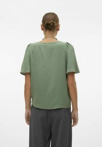 Blusa verde chiaro a maniche corte con finitura testurizzata, scollo rotondo e spalle leggermente imbottite, abbinata a pantaloni grigio scuro.