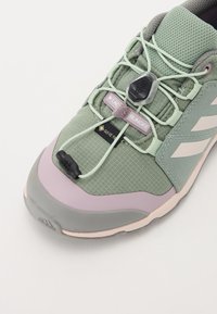Baskets de sport synthétiques vertes avec une tige en tissu texturé, un système de lacets élastiques et un col rembourré avec des accents gris et lavande.