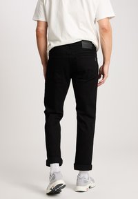 Zwarte jeans met een rechte pasvorm, opgerolde manchetten en een leren label op de taille. De broek heeft een fijne textuur en minimale details.