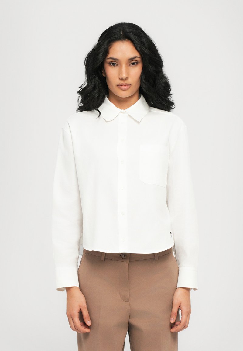 Chemise blanche cintrée à boutons, avec un col et une poche poitrine unique, assortie à un pantalon beige clair. Tissu lisse et léger.
