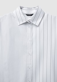 Camicia bianca con righe verticali nere, design con bottoni, colletto a punte e texture liscia. Etichetta del produttore visibile all'interno del colletto.