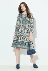 Mujer vistiendo un vestido paisley de largo hasta la rodilla con patrones en azul, negro y beige, sosteniendo un bolso azul y llevando mocasines azules.