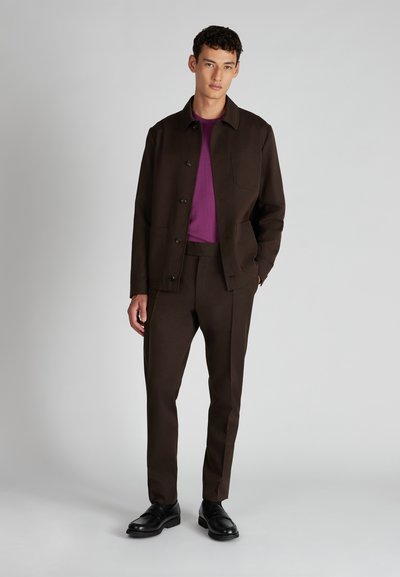 Brązowa kurtka z guzikami i kieszenią na piersi, zestawiona z dopasowanymi brązowymi spodniami. Pod spodem fioletowa koszula. Czarne buty dopełniają strój.