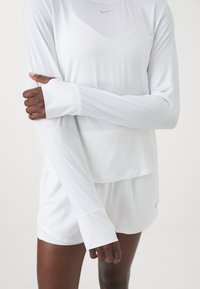 White long-sleeve athletic top with thumbholes, smooth fabric, round neckline, and Nike logo. Paired with white shorts.