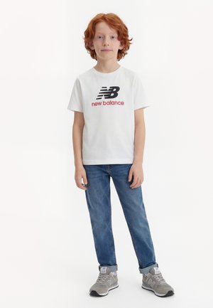 Niño con cabello rojo que lleva una camiseta blanca de New Balance, pantalones vaqueros azules y zapatillas grises de New Balance, de pie contra un fondo blanco.